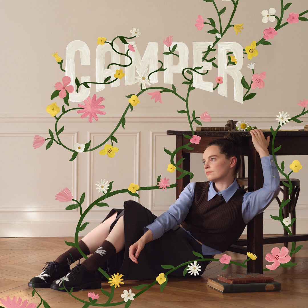 Emma Portner leans against a chair in black floral shoes as illustrated flowers grow across the room in CAMPER's Fall/Winter 2026 campaign.