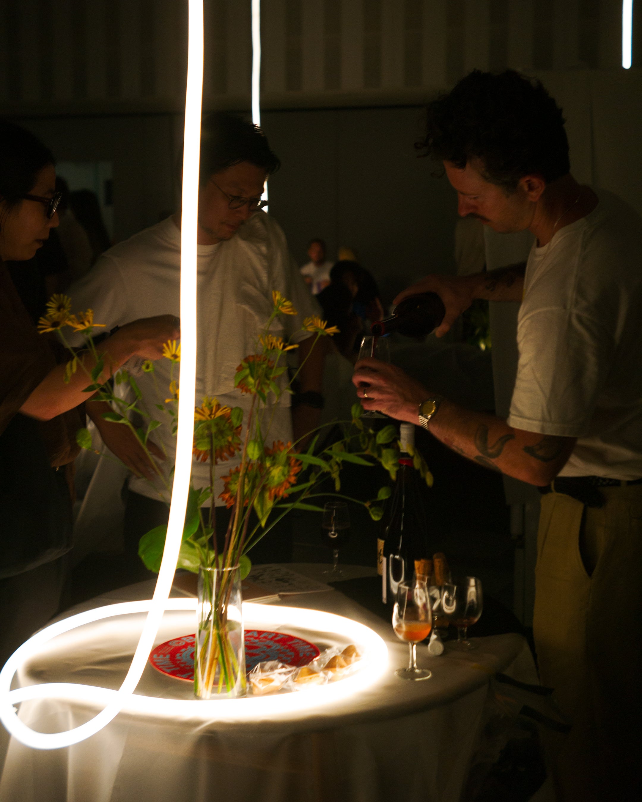 People pouring wine beside yellow and orange flowers at tables draped in white cloth, with tubular lighting